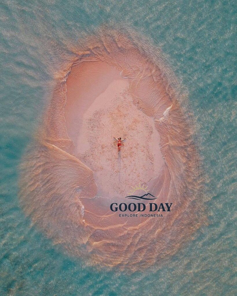 A breathtaking aerial view of Gili Pasir Lombok, featuring a tiny pink-tinted sandbar emerging from crystal-clear turquoise waters. A traveler lies at the center of the sandbank, surrounded by naturally sculpted tidal patterns, showcasing one of Lombok’s most unique and photogenic hidden island destinations.