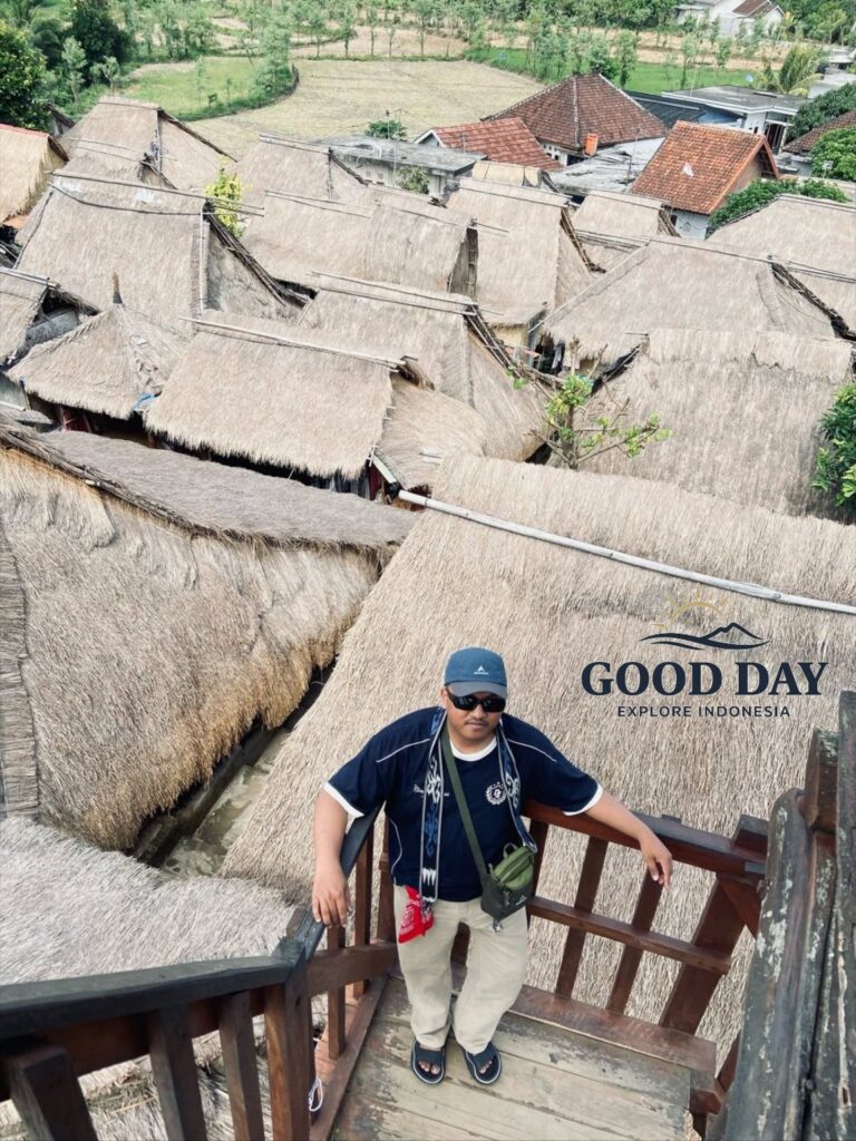 Visitor standing on a traditional wooden viewing platform overlooking the iconic thatched-roof houses of Sade Traditional Village in Lombok, Indonesia. The Sasak cultural village showcases authentic indigenous architecture, local heritage, and rural village life.