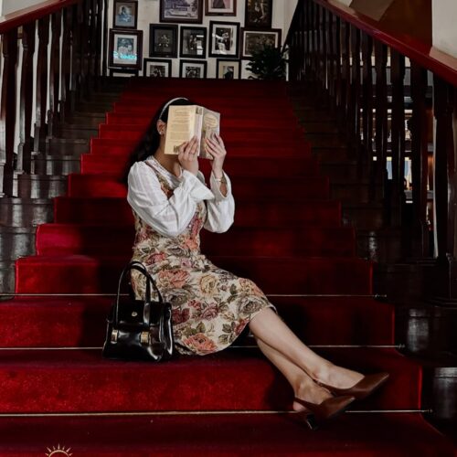 Traveler reading a book on the iconic red-carpet staircase inside Café Batavia, Jakarta. Surrounded by vintage photographs, polished wooden railings, and elegant colonial-era interiors, the scene captures the timeless charm and heritage atmosphere of Jakarta's historic Old Town.