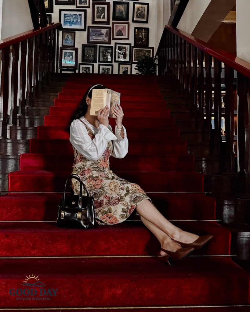 Traveler reading a book on the iconic red-carpet staircase inside Café Batavia, Jakarta. Surrounded by vintage photographs, polished wooden railings, and elegant colonial-era interiors, the scene captures the timeless charm and heritage atmosphere of Jakarta's historic Old Town.