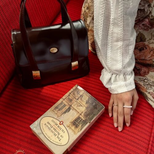Vintage-inspired still life inside Café Batavia Jakarta featuring a classic leather handbag, a copy of The Hunchback of Notre-Dame, and elegant period-style attire resting on the café's iconic red-upholstered seating. The scene captures the timeless charm and colonial atmosphere of Jakarta's historic Old Town.