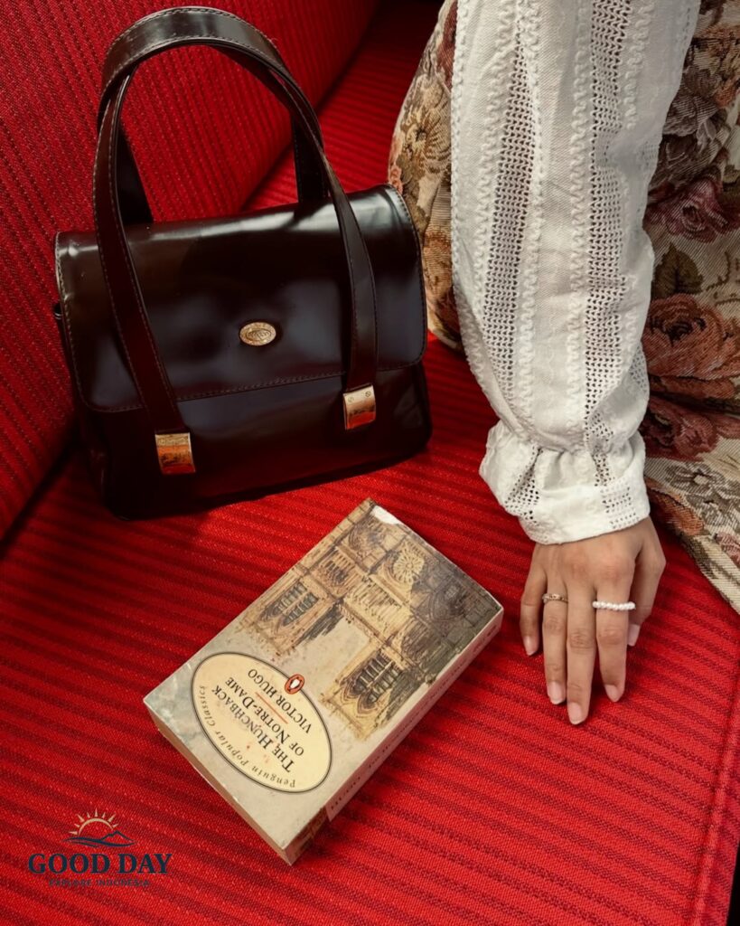 Vintage-inspired still life inside Café Batavia Jakarta featuring a classic leather handbag, a copy of The Hunchback of Notre-Dame, and elegant period-style attire resting on the café's iconic red-upholstered seating. The scene captures the timeless charm and colonial atmosphere of Jakarta's historic Old Town.