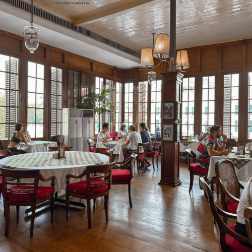 Elegant dining room inside Café Batavia Jakarta featuring colonial-era architecture, polished wooden floors, vintage furnishings, red-cushioned chairs, and large shuttered windows overlooking the historic Kota Tua district. Guests enjoy a relaxed dining experience in one of Jakarta's most iconic heritage restaurants.