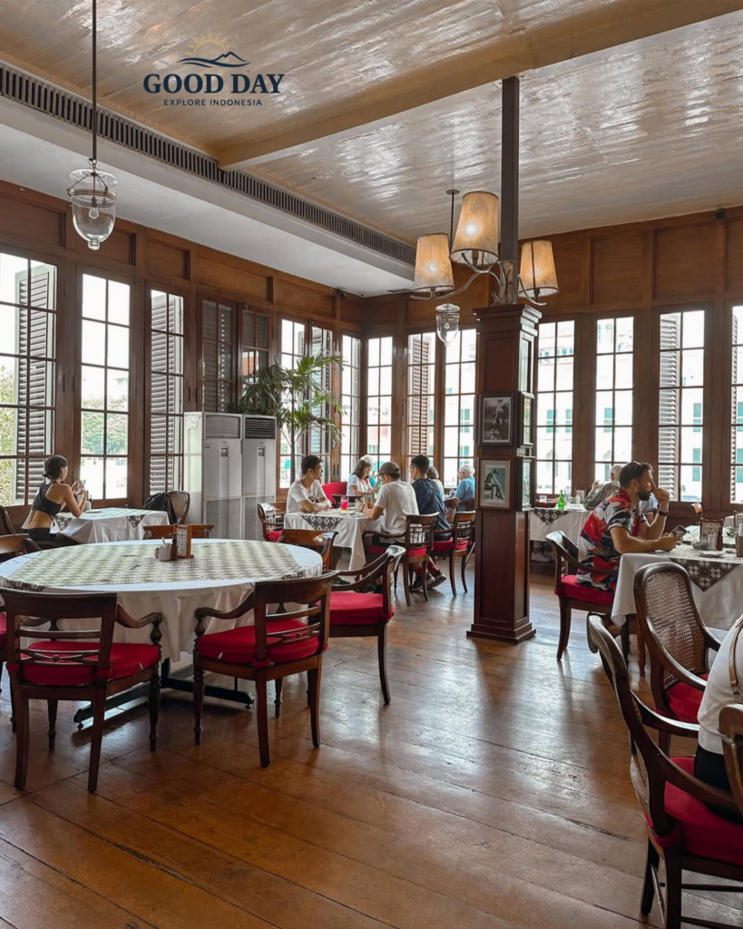 Elegant dining room inside Café Batavia Jakarta featuring colonial-era architecture, polished wooden floors, vintage furnishings, red-cushioned chairs, and large shuttered windows overlooking the historic Kota Tua district. Guests enjoy a relaxed dining experience in one of Jakarta's most iconic heritage restaurants.