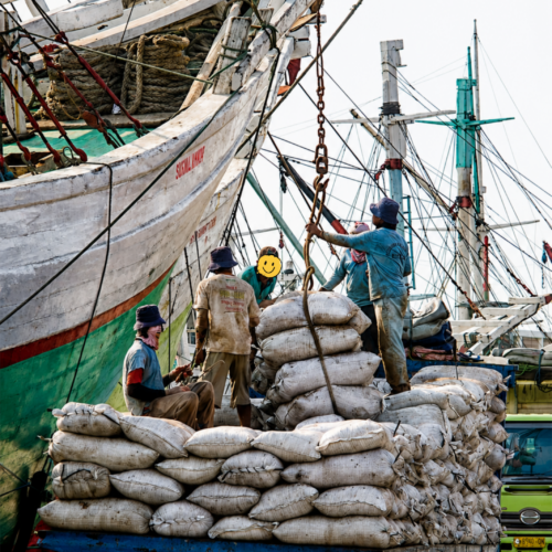 Cargo workers loading and unloading goods at Sunda Kelapa Harbour in Jakarta, Indonesia. Traditional wooden phinisi ships are docked alongside the port while laborers handle sacks of cargo, showcasing the daily maritime trade activities that have sustained Jakarta's historic harbor for centuries.