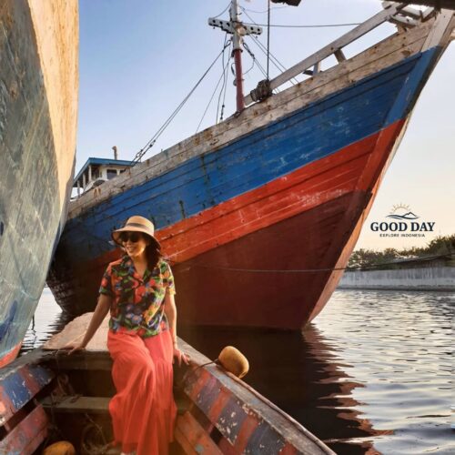 Traveler sitting on a traditional wooden boat at Sunda Kelapa Harbour, Jakarta, surrounded by colorful Indonesian phinisi ships during golden hour. The historic harbor showcases Jakarta's maritime heritage with calm waters, authentic wooden vessels, and a warm sunset atmosphere.