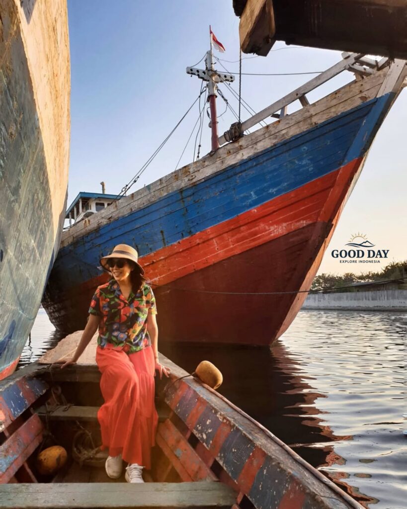 Traveler sitting on a traditional wooden boat at Sunda Kelapa Harbour, Jakarta, surrounded by colorful Indonesian phinisi ships during golden hour. The historic harbor showcases Jakarta's maritime heritage with calm waters, authentic wooden vessels, and a warm sunset atmosphere.