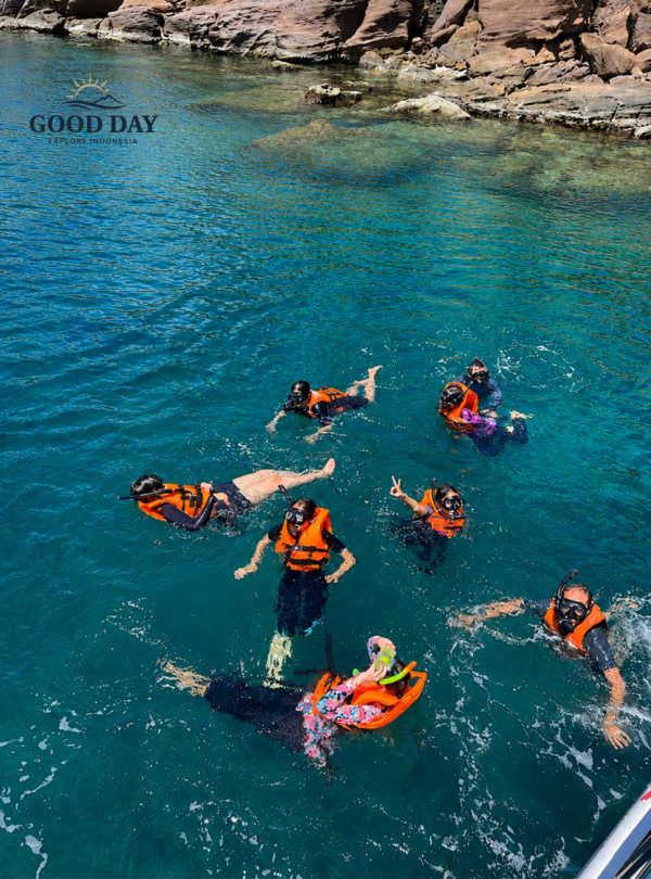 Group of tourists snorkeling in the crystal-clear turquoise waters of Bunaken Marine Park, Manado, North Sulawesi. Visitors wearing life jackets and snorkeling masks enjoy a marine adventure near a rocky coastline surrounded by pristine tropical waters.