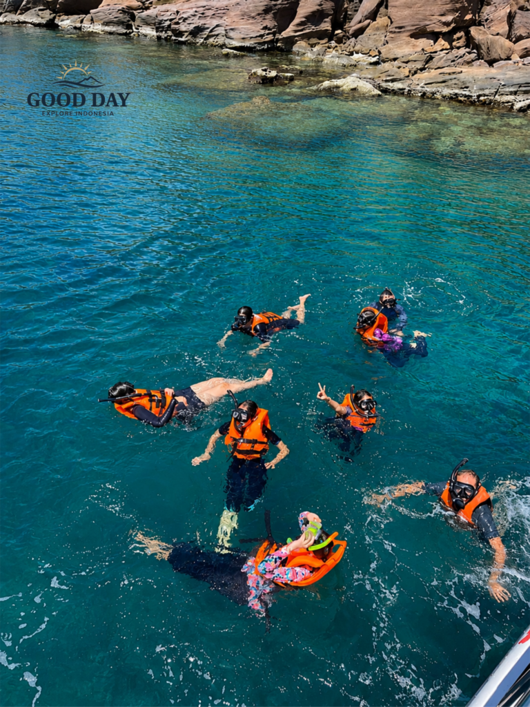 Group of tourists snorkeling in the crystal-clear turquoise waters of Bunaken Marine Park, Manado, North Sulawesi. Visitors wearing life jackets and snorkeling masks enjoy a marine adventure near a rocky coastline surrounded by pristine tropical waters.