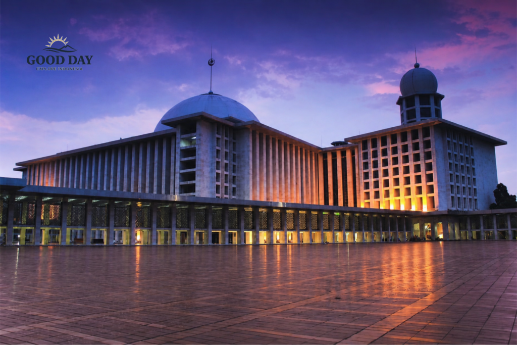 Masjid Istiqlal Jakarta illuminated at dusk with glowing exterior lights, reflecting on the expansive courtyard under a dramatic purple and blue evening sky. The grand mosque's iconic dome and modern Islamic architecture stand prominently as one of Indonesia's most important religious landmarks.