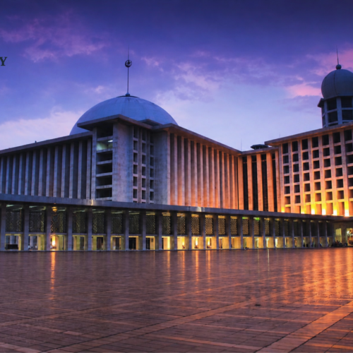 Masjid Istiqlal Jakarta illuminated at dusk with glowing exterior lights, reflecting on the expansive courtyard under a dramatic purple and blue evening sky. The grand mosque's iconic dome and modern Islamic architecture stand prominently as one of Indonesia's most important religious landmarks.
