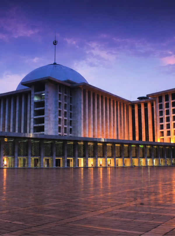 Masjid Istiqlal Jakarta illuminated at dusk with glowing exterior lights, reflecting on the expansive courtyard under a dramatic purple and blue evening sky. The grand mosque's iconic dome and modern Islamic architecture stand prominently as one of Indonesia's most important religious landmarks.