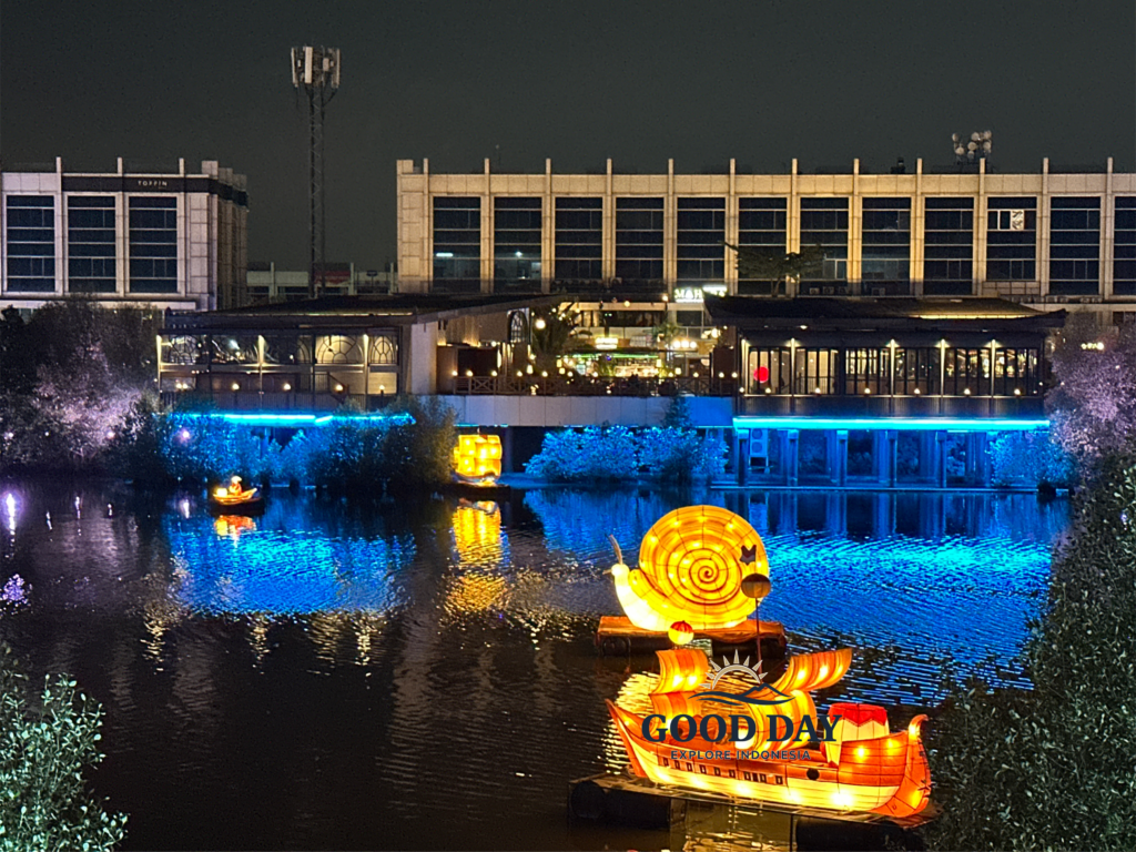 Colorful illuminated lantern installations floating on a lake at PIK Jakarta during the evening, featuring a glowing snail-shaped lantern boat reflected on calm water, surrounded by vibrant lights, restaurants, and modern waterfront scenery.