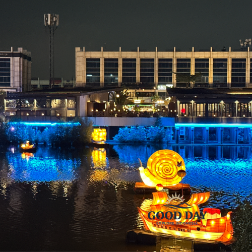 Colorful illuminated lantern installations floating on a lake at PIK Jakarta during the evening, featuring a glowing snail-shaped lantern boat reflected on calm water, surrounded by vibrant lights, restaurants, and modern waterfront scenery.