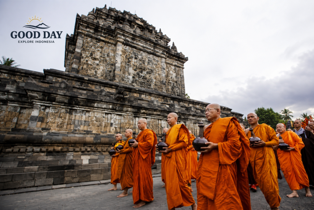 Buddhist monks in bright orange robes participate in a sacred religious procession at a historic temple in Yogyakarta, Indonesia. The monks walk barefoot while carrying ceremonial alms bowls, with ancient stone temple architecture and intricate relief carvings forming a dramatic cultural backdrop.