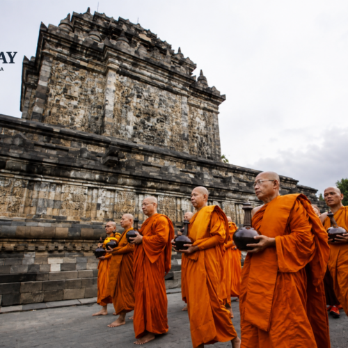 Buddhist monks in bright orange robes participate in a sacred religious procession at a historic temple in Yogyakarta, Indonesia. The monks walk barefoot while carrying ceremonial alms bowls, with ancient stone temple architecture and intricate relief carvings forming a dramatic cultural backdrop.