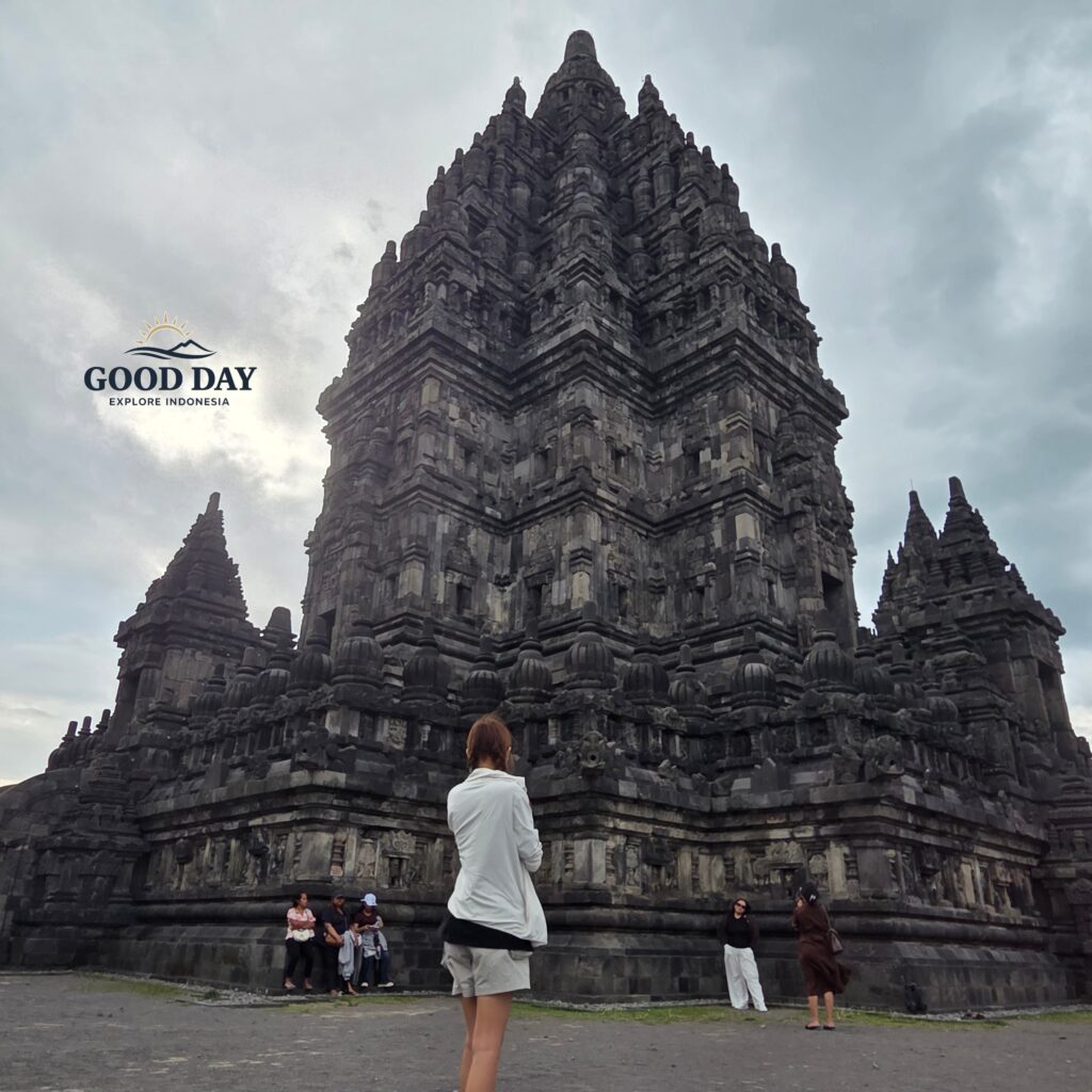 A traveler standing in front of the majestic Prambanan Temple in Yogyakarta, Indonesia, surrounded by towering ancient Hindu stone architecture under a dramatic cloudy sky. The iconic UNESCO World Heritage site showcases intricate carvings and centuries-old Javanese craftsmanship.
