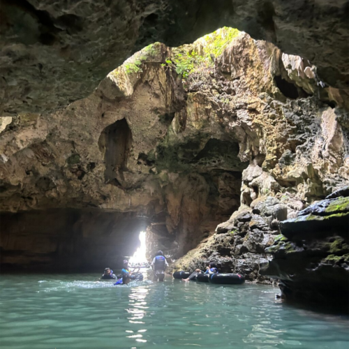 Visitors exploring the underground river inside Jomblang Cave in Yogyakarta, Indonesia, surrounded by towering limestone formations, crystal-clear water, and natural light entering through the cave opening.