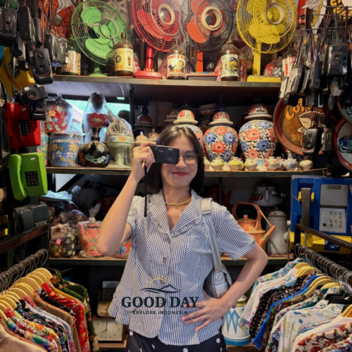 Visitor exploring a traditional market in Yogyakarta, Indonesia, standing among vintage collectibles, colorful batik clothing, antique fans, retro telephones, and classic decorative items inside a unique cultural souvenir shop.