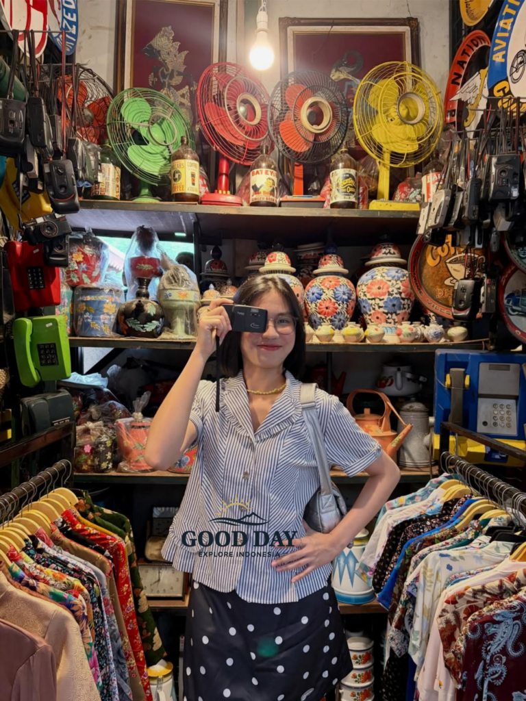 Visitor exploring a traditional market in Yogyakarta, Indonesia, standing among vintage collectibles, colorful batik clothing, antique fans, retro telephones, and classic decorative items inside a unique cultural souvenir shop.