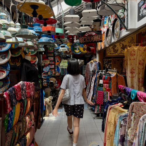 Visitor walking through a vibrant traditional market in Yogyakarta, Indonesia, surrounded by colorful batik fabrics, vintage enamel lamps, retro signs, and unique local handicrafts in a bustling cultural shopping alley.