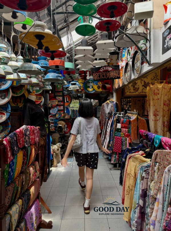 Visitor walking through a vibrant traditional market in Yogyakarta, Indonesia, surrounded by colorful batik fabrics, vintage enamel lamps, retro signs, and unique local handicrafts in a bustling cultural shopping alley.