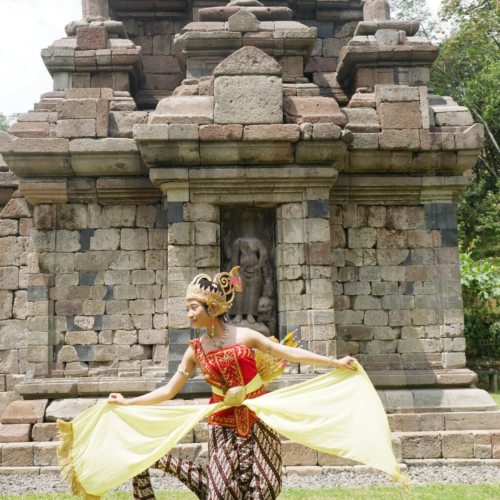 Traditional Javanese dancer performing a cultural dance in front of Selogriyo Temple, Yogyakarta, wearing colorful traditional attire with a historic ancient Hindu temple as the backdrop.