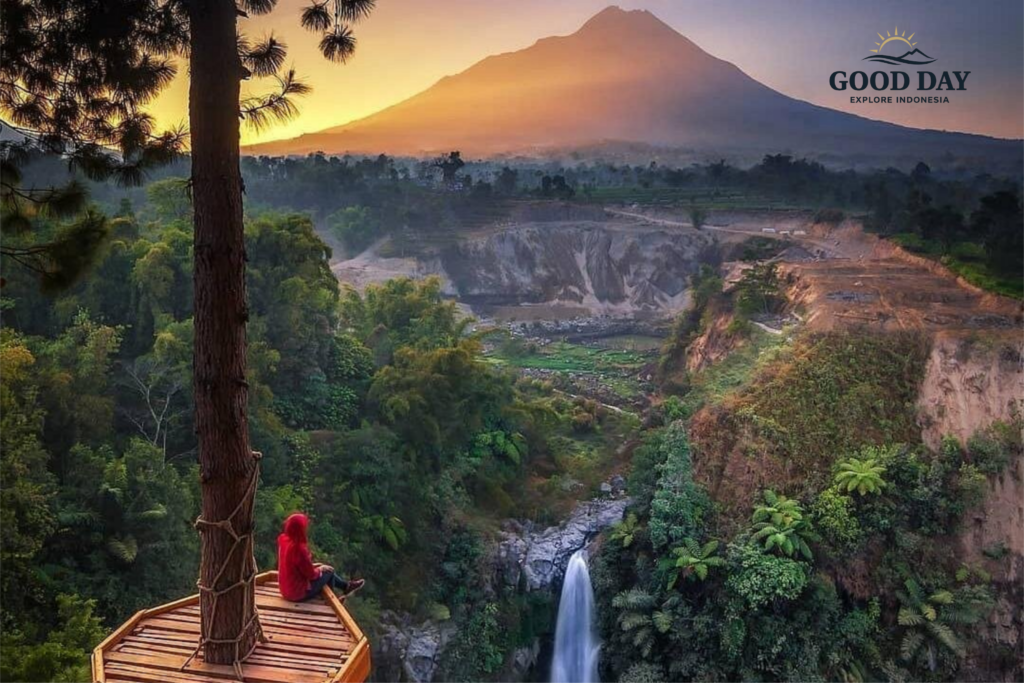 Traveler sitting on a wooden viewing platform overlooking Kedung Kayang Waterfall at sunrise in Yogyakarta, Indonesia, with lush green forest, dramatic canyon cliffs, and the majestic silhouette of Mount Merapi in the background.