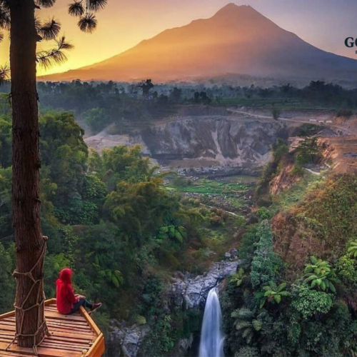 Traveler sitting on a wooden viewing platform overlooking Kedung Kayang Waterfall at sunrise in Yogyakarta, Indonesia, with lush green forest, dramatic canyon cliffs, and the majestic silhouette of Mount Merapi in the background.