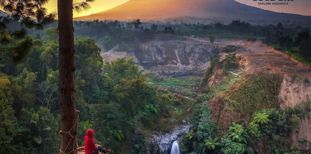 Traveler sitting on a wooden viewing platform overlooking Kedung Kayang Waterfall at sunrise in Yogyakarta, Indonesia, with lush green forest, dramatic canyon cliffs, and the majestic silhouette of Mount Merapi in the background.