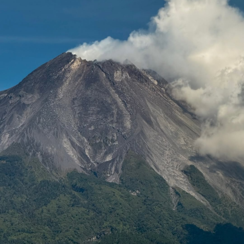 Close-up view of Mount Merapi volcano rising above the green landscapes of Yogyakarta, Indonesia, with dramatic volcanic slopes, rugged terrain, and clouds drifting around the summit.