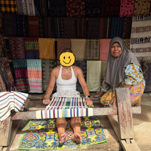 Visitor learning traditional Sasak weaving from a local artisan in Sade Traditional Village, Lombok, Indonesia, surrounded by colorful handwoven textiles and authentic cultural craftsmanship.
