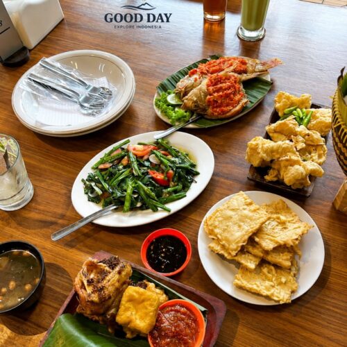 Traditional Sundanese meal in Bandung featuring spicy grilled fish with sambal, stir-fried water spinach, fried tofu, crispy tempeh, fresh vegetables, rice wrapped in banana leaves, and traditional Indonesian side dishes served on a wooden table.