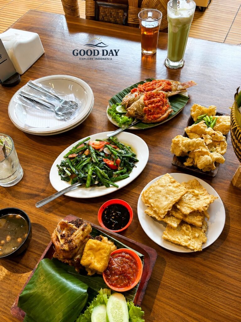 Traditional Sundanese meal in Bandung featuring spicy grilled fish with sambal, stir-fried water spinach, fried tofu, crispy tempeh, fresh vegetables, rice wrapped in banana leaves, and traditional Indonesian side dishes served on a wooden table.