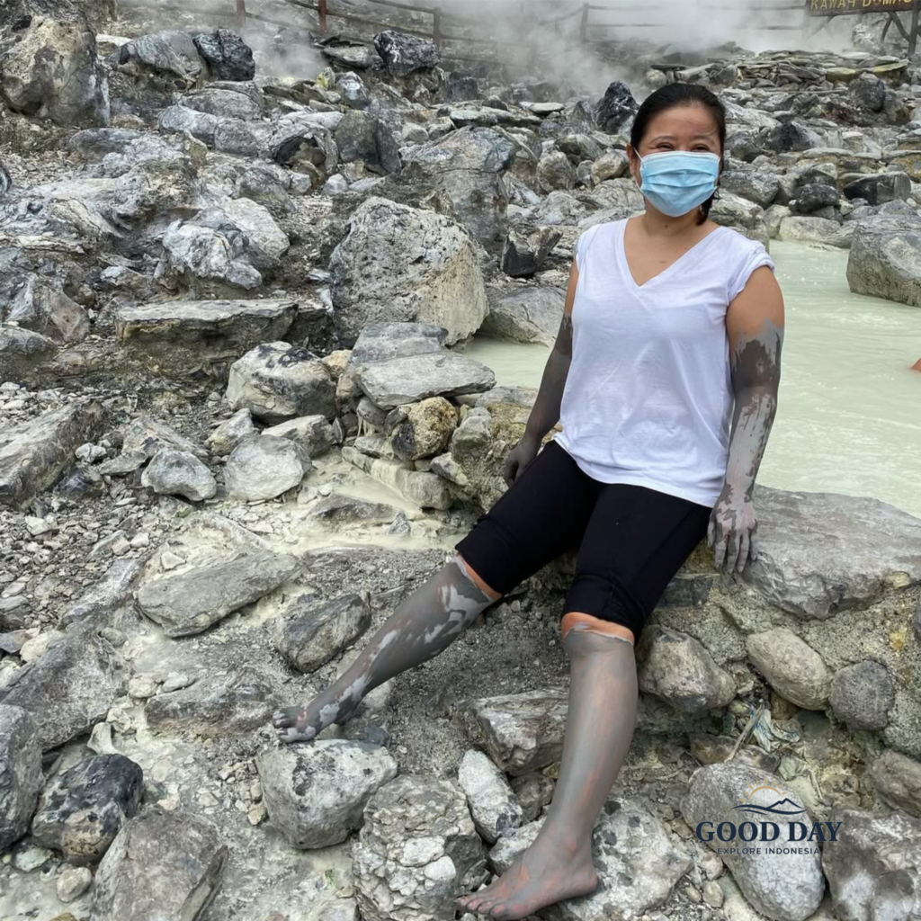Visitor covered in natural volcanic mud at Tangkuban Perahu, Bandung, sitting among steaming geothermal rocks and mineral-rich hot spring pools in West Java, Indonesia.