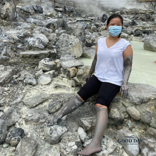 Visitor covered in natural volcanic mud at Tangkuban Perahu, Bandung, sitting among steaming geothermal rocks and mineral-rich hot spring pools in West Java, Indonesia.
