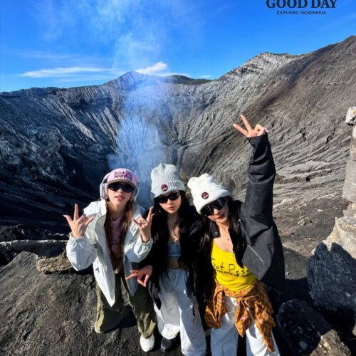 Three travelers posing at the rim of Mount Bromo Crater with volcanic smoke rising from the active crater in the background. The dramatic volcanic landscape of Bromo Tengger Semeru National Park, East Java, Indonesia, showcases rugged ash-covered slopes and breathtaking crater views.