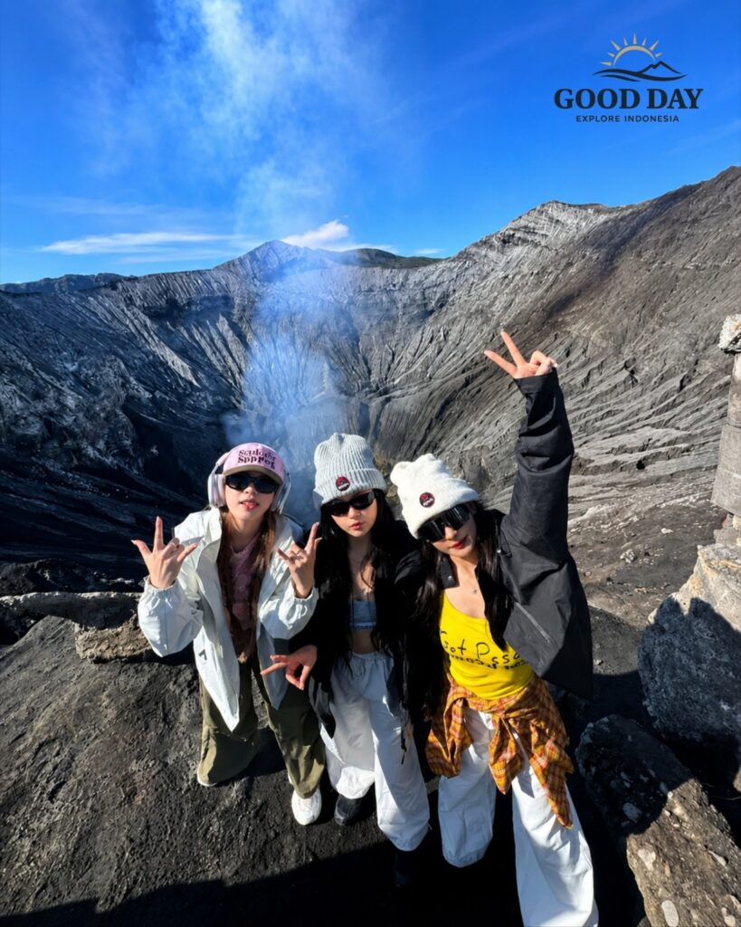 Three travelers posing at the rim of Mount Bromo Crater with volcanic smoke rising from the active crater in the background. The dramatic volcanic landscape of Bromo Tengger Semeru National Park, East Java, Indonesia, showcases rugged ash-covered slopes and breathtaking crater views.