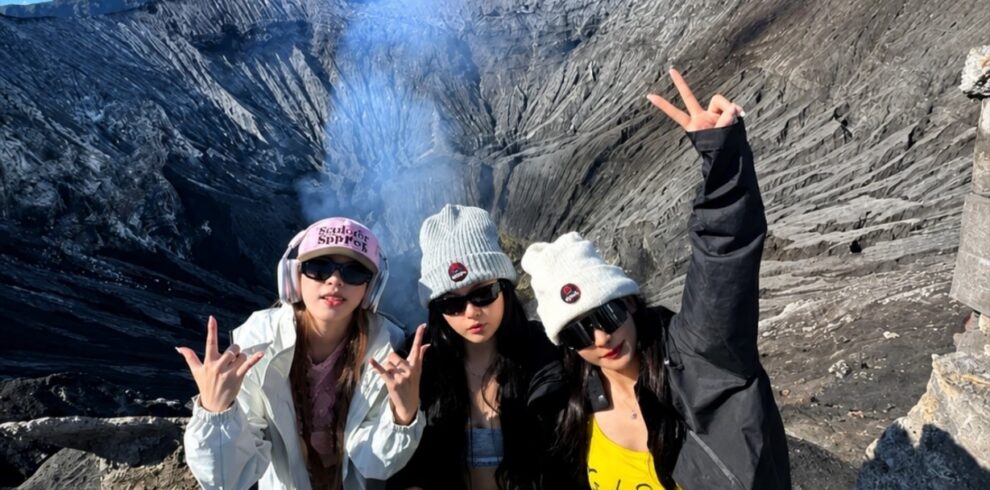 Three travelers posing at the rim of Mount Bromo Crater with volcanic smoke rising from the active crater in the background. The dramatic volcanic landscape of Bromo Tengger Semeru National Park, East Java, Indonesia, showcases rugged ash-covered slopes and breathtaking crater views.