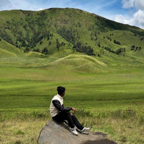 Traveler sitting on a rock overlooking the rolling green hills of Teletubbies Hill in Mount Bromo, East Java, Indonesia, surrounded by vast grasslands and scenic volcanic landscapes.