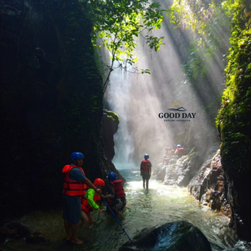 Adventure travelers exploring the hidden canyon of Curug Naga in Bogor, West Java, walking through a crystal-clear river beneath dramatic sun rays filtering through a lush tropical rainforest and waterfall gorge.