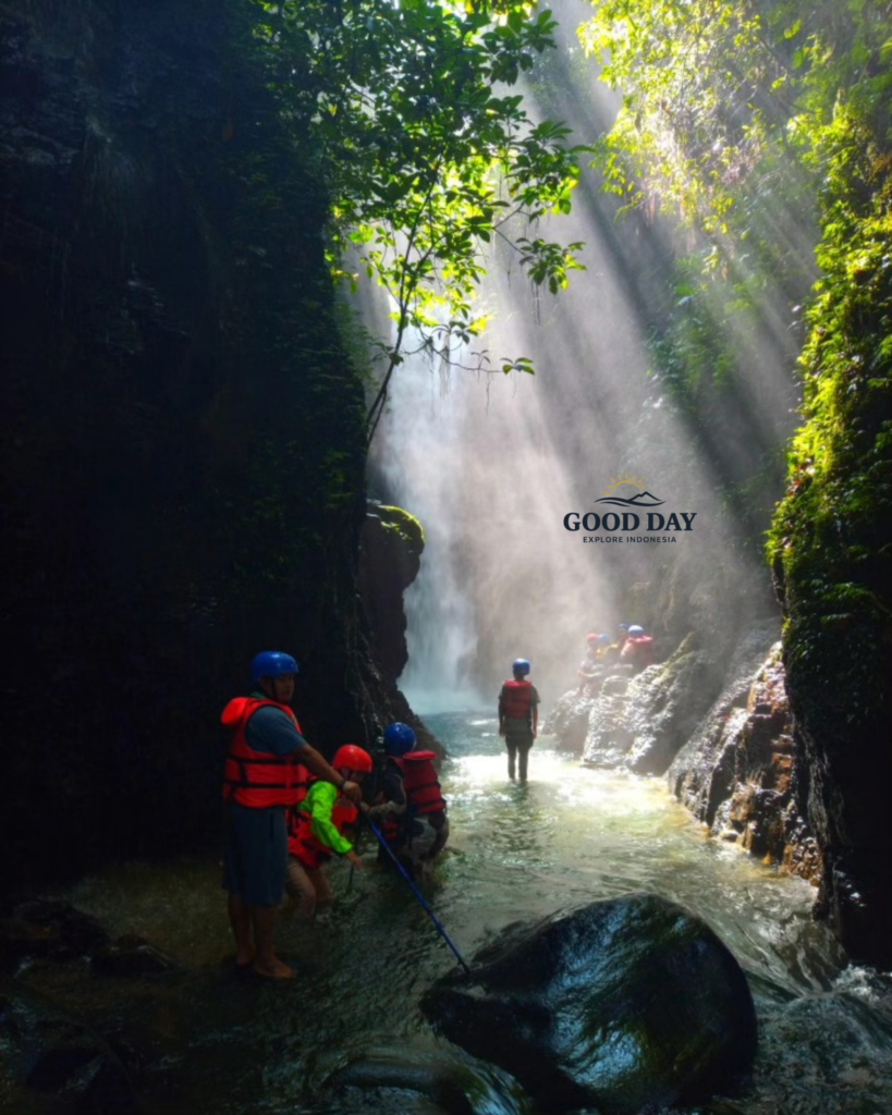 Adventure travelers exploring the hidden canyon of Curug Naga in Bogor, West Java, walking through a crystal-clear river beneath dramatic sun rays filtering through a lush tropical rainforest and waterfall gorge.