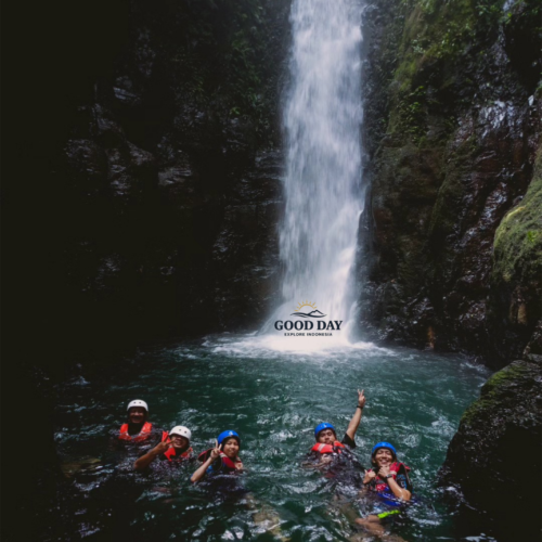 Visitors swimming beneath Curug Naga waterfall in Bogor, West Java, enjoying a body rafting adventure in a crystal-clear natural pool surrounded by towering canyon walls and lush tropical rainforest.