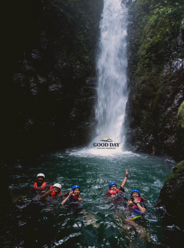 Visitors swimming beneath Curug Naga waterfall in Bogor, West Java, enjoying a body rafting adventure in a crystal-clear natural pool surrounded by towering canyon walls and lush tropical rainforest.