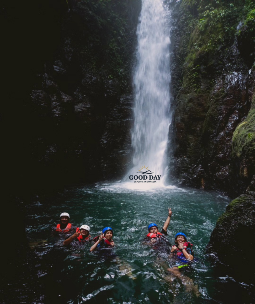 Visitors swimming beneath Curug Naga waterfall in Bogor, West Java, enjoying a body rafting adventure in a crystal-clear natural pool surrounded by towering canyon walls and lush tropical rainforest.