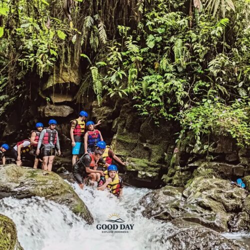 Group of adventure travelers participating in a body rafting experience at Curug Naga, Bogor, navigating a rocky river surrounded by lush tropical rainforest, moss-covered cliffs, and natural waterfalls in West Java, Indonesia.