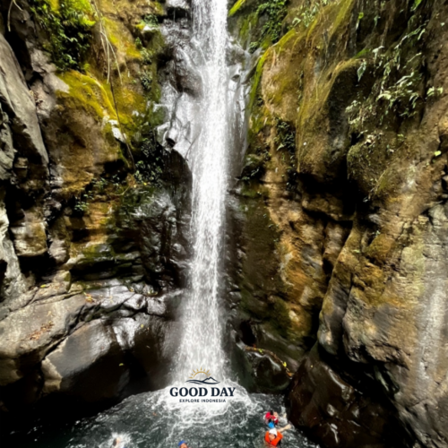 Visitors enjoying a body rafting adventure beneath Curug Naga waterfall in Bogor, West Java, surrounded by towering rock cliffs, moss-covered canyon walls, and crystal-clear natural pools.