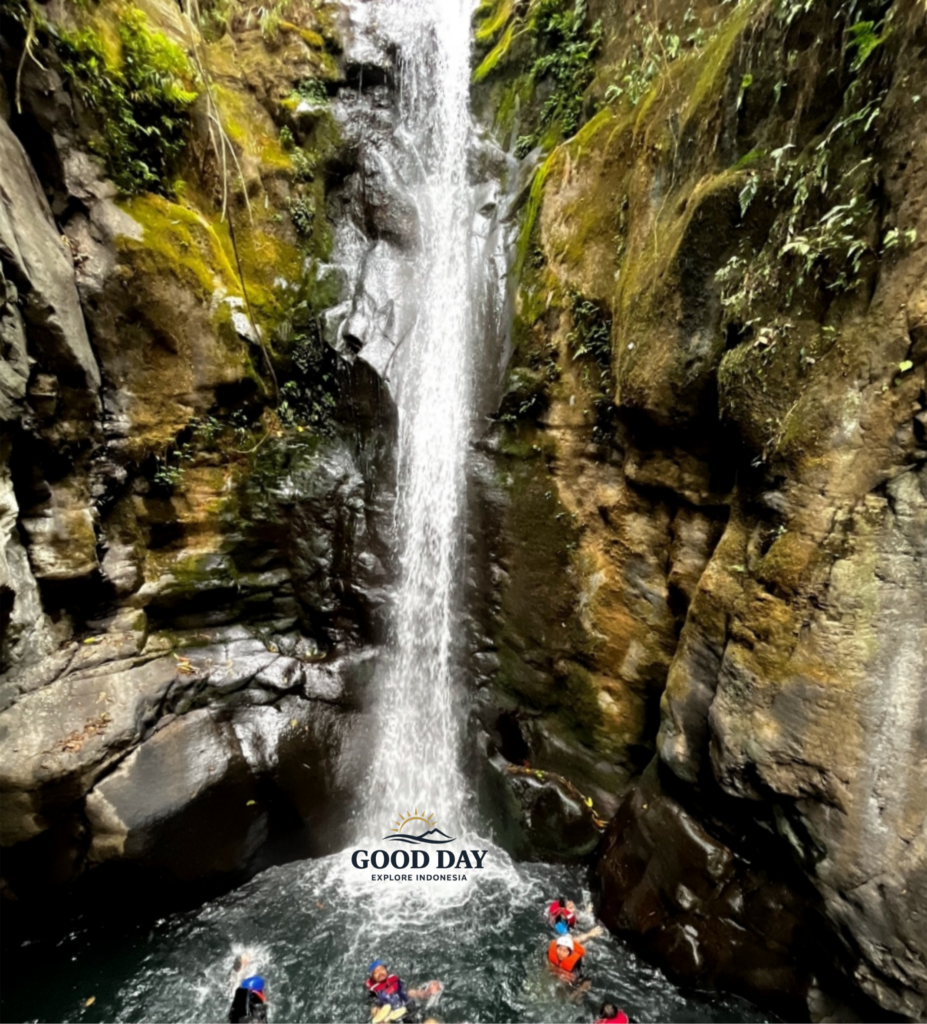 Visitors enjoying a body rafting adventure beneath Curug Naga waterfall in Bogor, West Java, surrounded by towering rock cliffs, moss-covered canyon walls, and crystal-clear natural pools.
