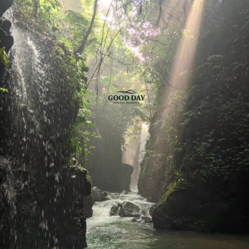 Sunlight streams through the lush rainforest canopy at Curug Naga, Bogor, illuminating a scenic waterfall canyon with crystal-clear river water, moss-covered cliffs, and tropical vegetation in West Java, Indonesia.