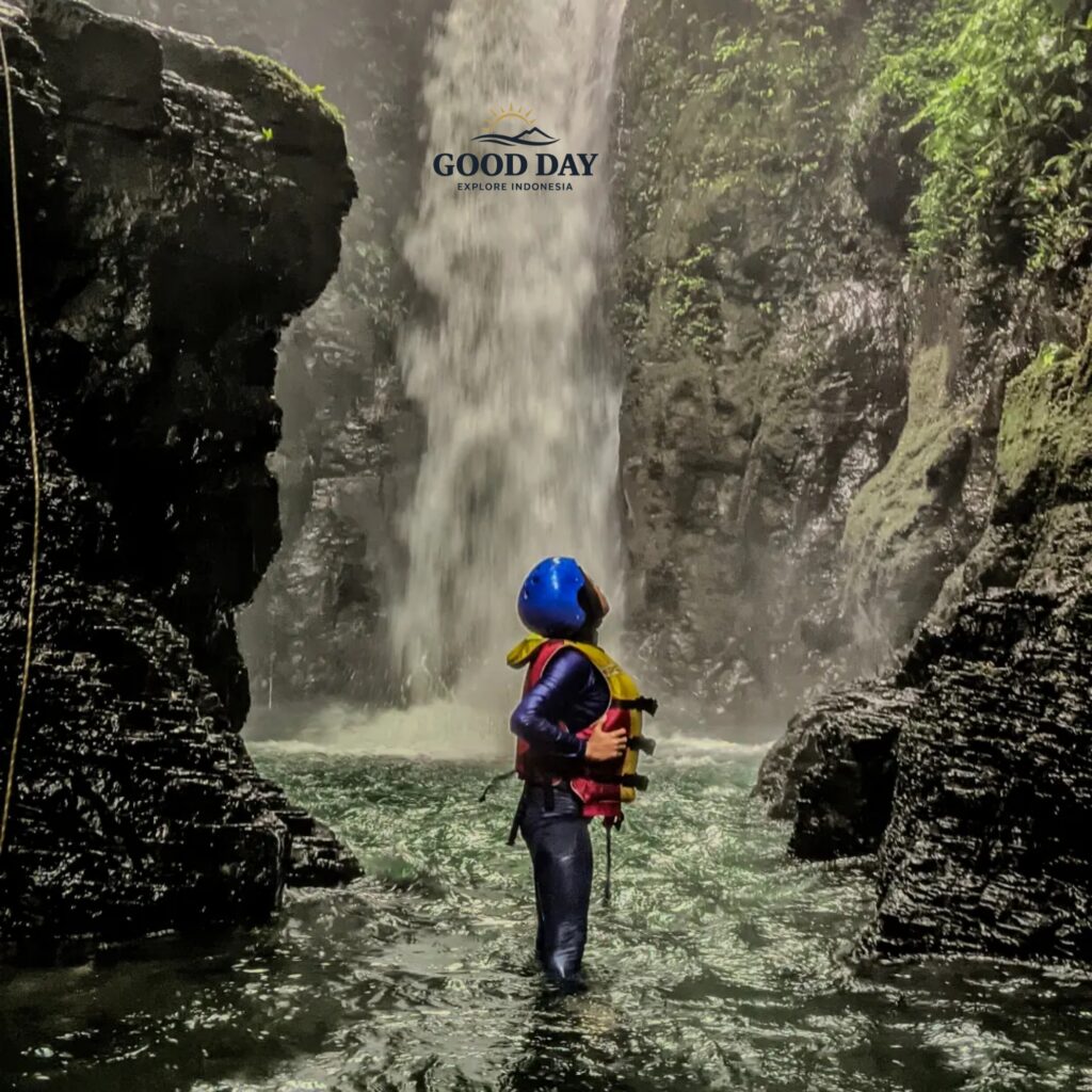 Solo adventurer standing in the water beneath Curug Naga waterfall in Bogor, West Java, wearing safety gear while surrounded by towering canyon walls, cascading water, and lush tropical rainforest.